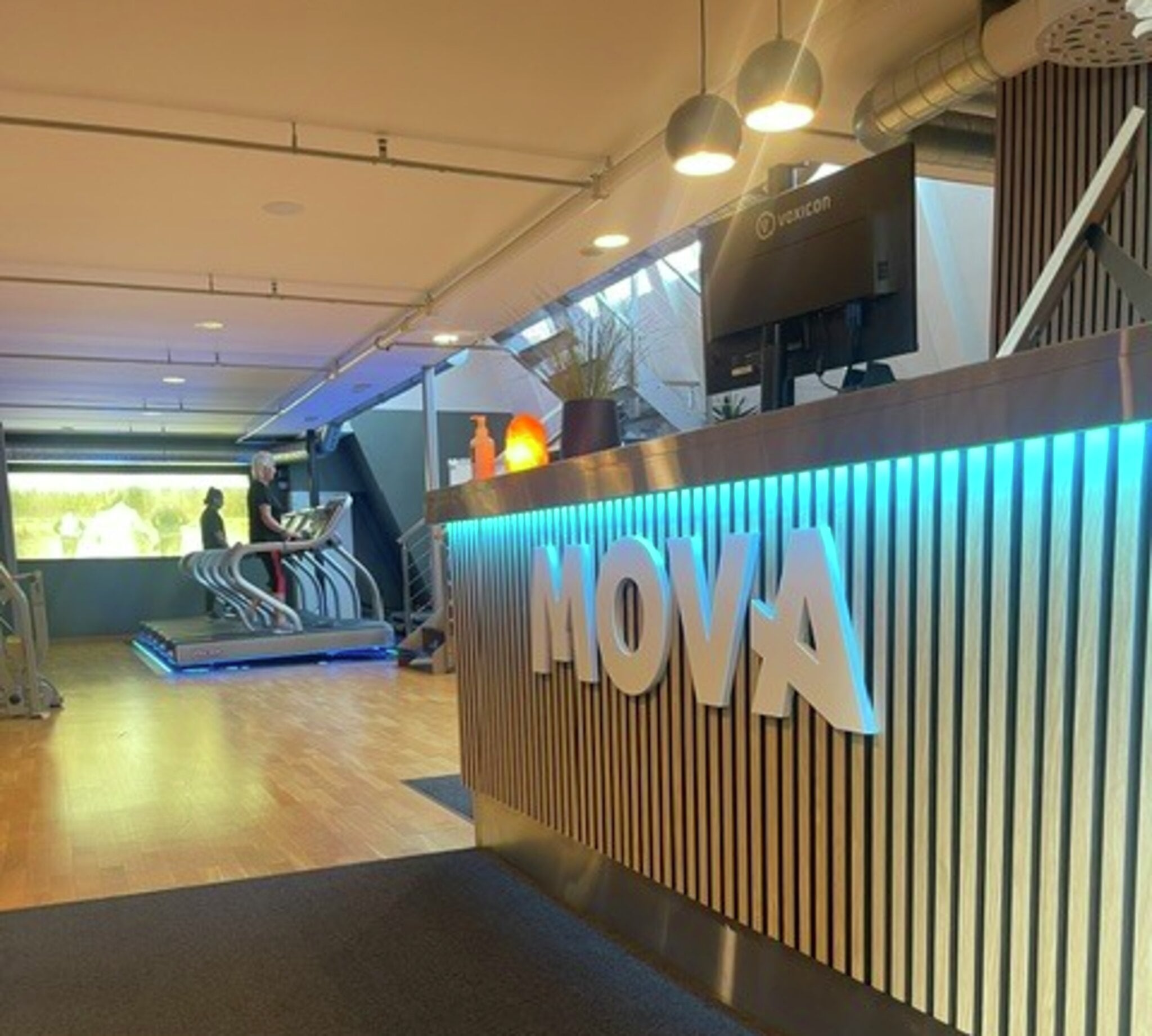 Resepsjonsdisk i trespiler med MOVA-logo og blå LED-lys, med tredemøller og dagslys bak på MOVA Kristiansund. Resepsjonen på MOVA Kristiansund møter deg rett innenfor døren, med disk i trespiler, MOVA-logoen i hvitt og blå LED-lys under logoen. Innenfor åpner senteret seg mot kardio-avdelingen med tredemøller på lyst tregulv og store vinduer som slipper inn dagslys.