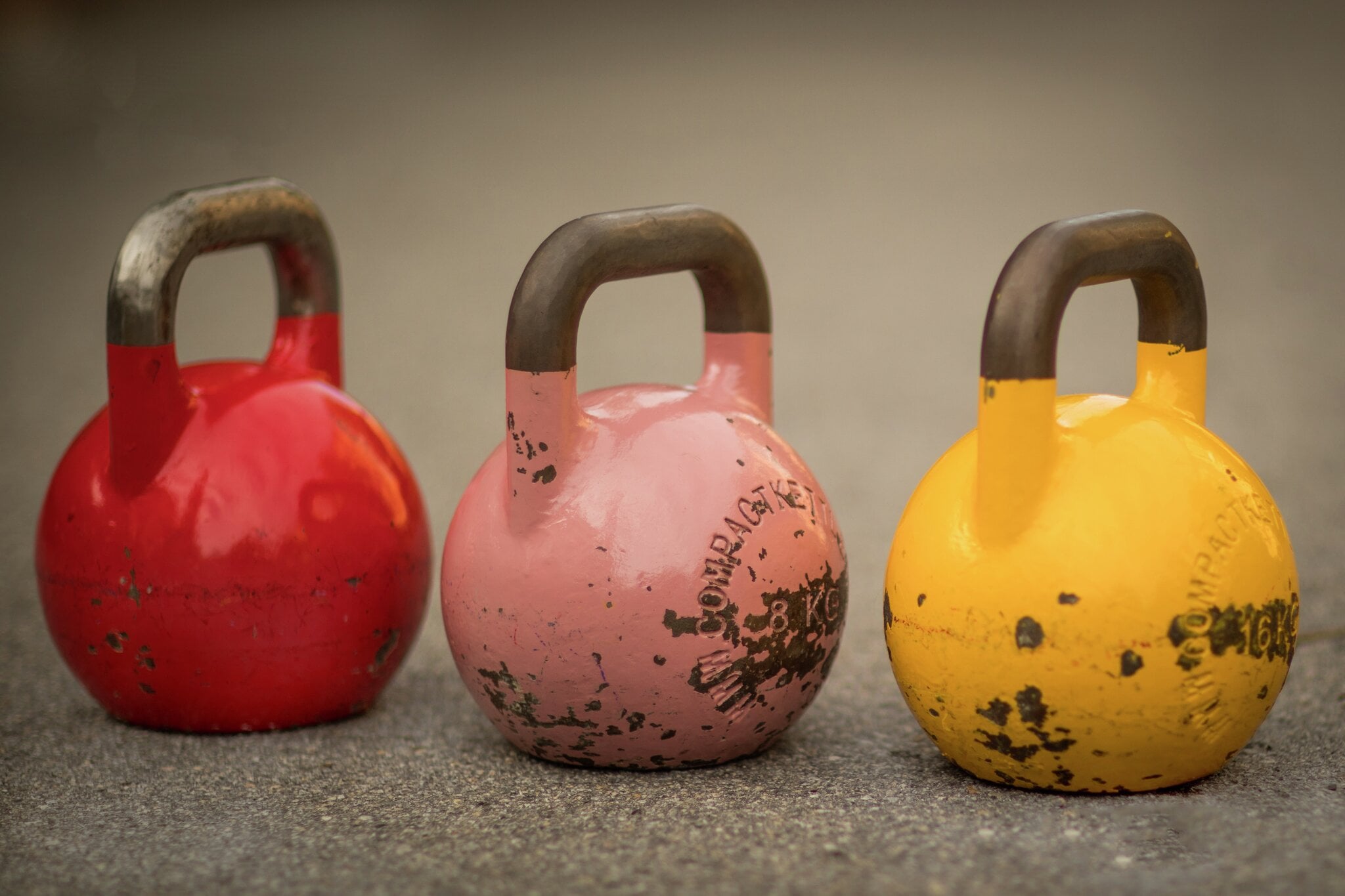 Rød, rosa og gul Compact-kettlebell står side ved side på betonggulv på MOVA Pindsle. Nærbilde av tre konkurranse-kettlebells i rødt, rosa og gult med tydelig patina mot et nøytralt betonggulv. De rene fargene fanger detaljen i frivektsonen og det velbrukte utstyret som står klart til funksjonelle økter på MOVA Pindsle.