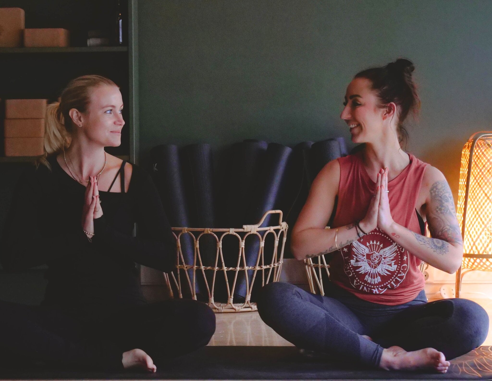 To kvinner sitter i skreddersittende namaste-positur i yogastudio på MOVA Pindsle. To kvinner sitter ansikt til ansikt i skreddersittende stilling med hendene samlet foran brystet i yogastudioet. Yogamatter er rullet sammen i en rotting-kurv bak, og det varme lyset fra lampen og mosegrønn vegg gir et rolig, hjemlig uttrykk.
