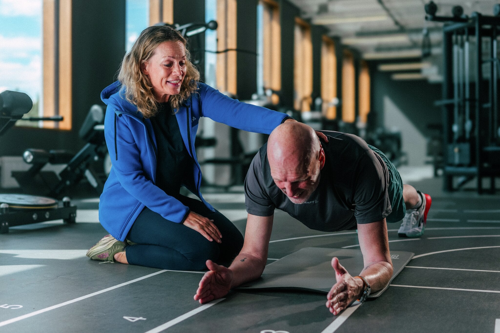Personlig trener veileder et mannlig medlem som tar planken på en matte på MOVA. En personlig trener sitter ved siden av et mannlig medlem som utfører planken på en treningsmatte. Treneren har en hånd på ryggen hans og veileder ham gjennom øvelsen på et treningssenter hos MOVA.