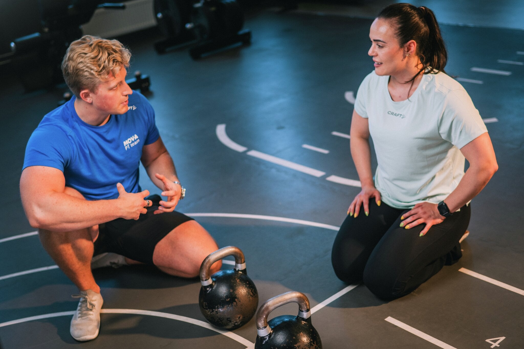 Personlig trener sitter på gulvet og snakker med en medlem ved to kettlebells, på MOVA. En personlig trener og en medlem sitter på gulvet i treningssalen og snakker sammen mellom øvingene. To kettlebells står foran dem. Personlig trening handler også om god dialog og planlegging av økten.