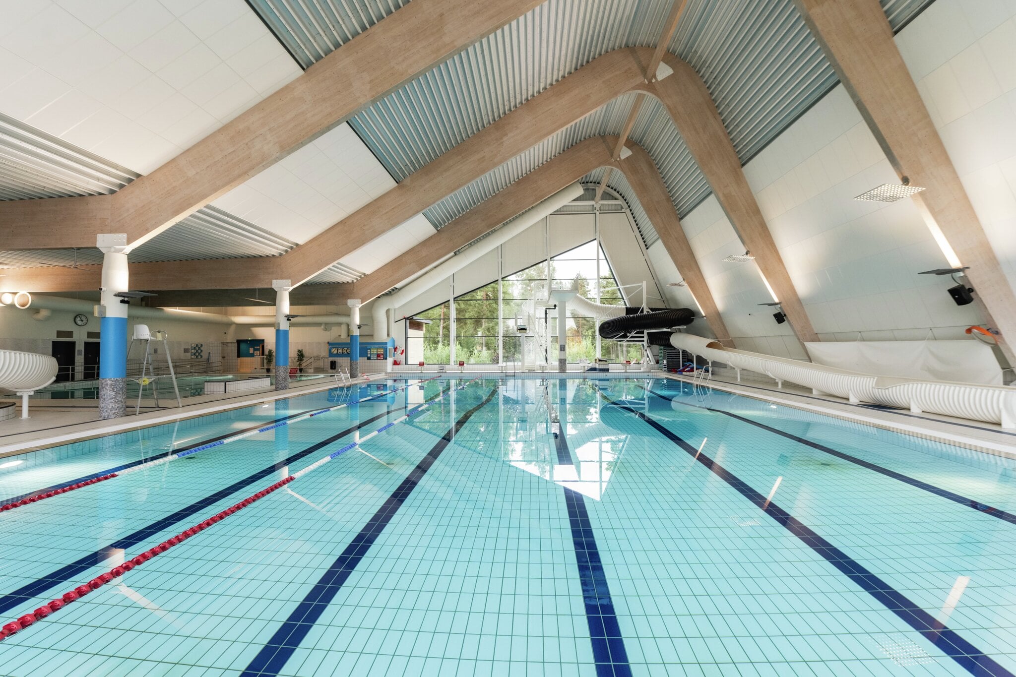 Svømmebaner, vannsklier og stupetårn i den store svømmehallen på MOVA Ringeriksbadet. Hovedbassenget strekker seg fra forgrunnen mot glassfasaden med stupetårn og vannsklier i enden. Banetau og bunnmarkeringer viser svømmebanene. Til venstre ligger et grunnere basseng med badevaktstol, under buede limtrebjelker.